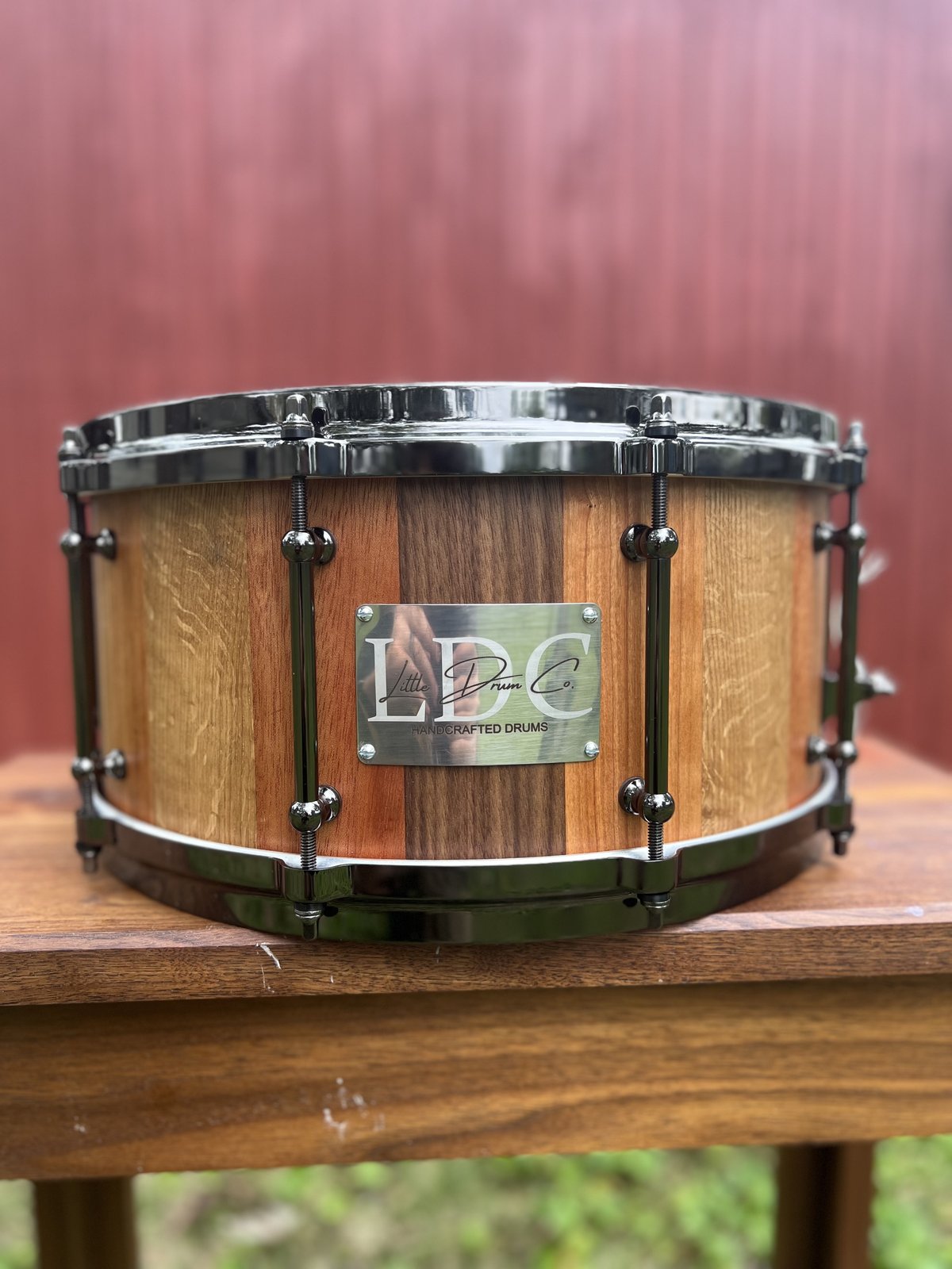 Stave snare drum built from alternating light and dark hardwoods, with black nickel tube lugs and hoops